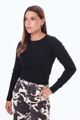 Remera de manga larga en color negro, confeccionada en tejido de punto acanalado. Presenta un cuello redondo con una pequeña abertura frontal cerrada por cuatro botones, ofreciendo un ajuste entallado al cuerpo.