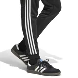 Pantalón deportivo Adidas modelo Tiro Esencial, color negro con las icónicas tres tiras blancas laterales y logo de la marca bordado en el muslo. Presenta corte ajustado, cintura elástica con cordón y puños acanalados en los tobillos.