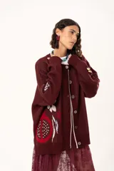 Saco de punto color vino con diseño holgado, cuello redondo y mangas largas. Presenta un gran bordado central en la espalda con forma de flor en tonos crema y rojo, acompañado de un pequeño bordado de sol y una frase bordada en la parte inferior.