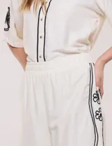 Conjunto de dos piezas negro compuesto por camisa de manga corta con cuello camisero y pantalón palazzo de tiro alto. Presenta detalles de bordados florales en contraste a lo largo de los laterales del pantalón y en las mangas.