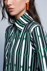 Camisa de corte holgado confeccionada en crepé fino, con estampado geométrico de rayas verticales en tonos verde, blanco y negro. Presenta cuello camisero de tamaño ligeramente ampliado, cierre frontal con botones y mangas largas con puños anchos.