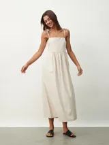 Vestido midi color beige claro, de corte amplio y fluido, con tirantes finos ajustables mediante lazos en los hombros, escote recto y prácticos bolsillos laterales.