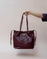 Cartera tipo tote de cuero color bordó, con diseño fruncido en los laterales y abertura central integrada como asa. Presenta una silueta flexible y espaciosa, ideal para uso diario.