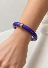 Pulsera rígida de resina en color violeta, con detalle central de aplique metálico dorado.