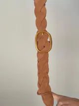 Cinturón trenzado de estilo artesanal confeccionado en material símil gamuza color tostado, con hebilla ovalada metálica en tono dorado.