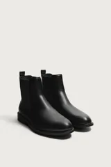 Bota estilo Chelsea de color negro, con parte superior lisa, paneles elásticos laterales y tirador trasero. Cuenta con horma redonda y suela con ligero tacón.