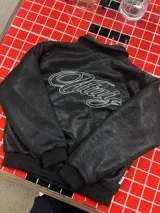 Campera de cuero sintético negro con diseño tipo bomber, cuello camisero y puños y ruedo elásticos. Presenta un bordado grande en la espalda con tipografía cursiva en color blanco.