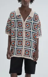 Camisa de manga corta, tejida a crochet con un patrón de cuadrados en colores beige, naranja y turquesa. Presenta cuello tipo polo y cierre frontal con botones.