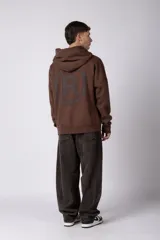 Buzo canguro color marrón con capucha, confeccionado en frisa de algodón. Presenta un diseño gráfico estampado en el frente con texto y logo circular, bolsillo tipo canguro y puños acanalados.