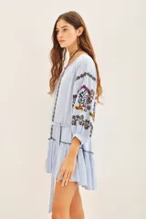 Vestido corto de bambula color celeste, con escote redondo, mangas tres cuartos abullonadas y falda con volado en el ruedo. Presenta bordados decorativos en el frente y las mangas, y cuenta con un cinturón de la misma tela para ajustar a la cintura.