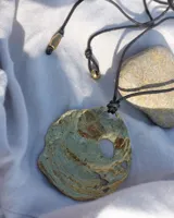 Collar artesanal con dije central de metal con textura orgánica irregular que evoca una ostra. El dije está montado sobre un cordón de algodón natural, permitiendo un uso versátil y regulable.