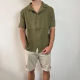 Camisa de hombre color verde oliva, de muselina con textura arrugada, manga corta, corte oversize, cuello cubano y cierre con botones. Presenta un pequeño logo bordado en el pecho.