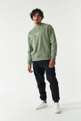 Buzo de felpa color verde oliva, con cuello redondo, puños y ruedo acanalados. Presenta un estampado frontal con texto en inglés y japonés junto al logo de la marca Mistral, y un detalle de etiqueta en la manga.