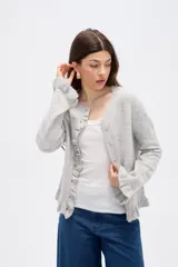 Saco de punto color gris, con cuello redondo, cierre frontal con botones y detalle de volados en la tapeta y el ruedo. Presenta mangas largas con puños acampanados.