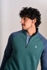 Buzo de cuello alto con cierre frontal parcial, diseño bicolor con cuerpo en verde y mangas raglán en azul. Presenta el logo de la marca bordado en el pecho.