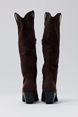 Bota de caña alta confeccionada en gamuza color chocolate, con diseño de estilo western, punta fina y taco cuadrado de altura media.
