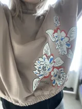 Buzo de algodón color tostado, de corte cropped y estilo oversize. Presenta cuello redondo, mangas largas con puños ajustados y elástico en la cintura. Destaca por una estampa exclusiva de flores en el frente y en una de las mangas.