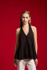 Blusa de algodón color beige con escote en V, cuello halter y cierre frontal con botones. Presenta un diseño ajustado en el torso con pinzas y pliegues en la parte inferior que generan una caída ligeramente acampanada.