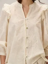 Camisa de color crema con bordados florales, cuello mao, cierre frontal con botones y mangas largas abullonadas con volados en los hombros.