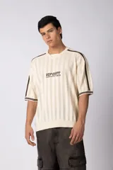 Remera de tejido de punto con textura calada, color crema con rayas verticales sutiles. Presenta cuello en V, mangas cortas con franjas laterales en color marrón oscuro y logo bordado en el centro del pecho.