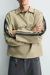 Sudadera de corte boxy fit color verde oliva, con cuello solapa y abertura frontal. Presenta mangas largas con bandas laterales negras a contraste y bordado frontal con texto en color negro.