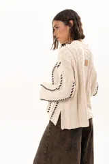 Buzo de tejido de punto color crudo con diseño de trenzas y costuras decorativas en color negro que recorren la prenda en forma de espiral. Presenta cuello redondo y mangas largas con terminación acampanada.