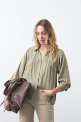 Camisa de color verde oliva con cuello clásico, cierre frontal de botones y diseño con tablas verticales en el frente. Presenta mangas largas con puños abotonados y un corte relajado.