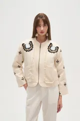 Campera estilo bomber color beige claro, con cuello redondo, cierre frontal y detalles de volados en los bordes. Presenta bordados florales en negro y blanco en la zona del pecho y bordados de tréboles negros en las mangas.
