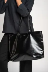 Bolso tipo tote de gran tamaño en cuero vegano color negro con acabado brillante. Cuenta con doble asa de hombro y placa metálica decorativa con logo en la parte frontal.