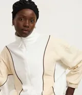 Campera deportiva de cuello alto con cierre frontal completo. Presenta un diseño bicolor en blanco y beige con líneas curvas en negro que definen la silueta. Confeccionada en una mezcla de poliéster, modal y elastano.