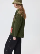 Campera tejido grueso y abrigado color verde militar, con forro de nylon interno, cuello estilo camisa en color negro y cierre frontal con botones. Cuenta con cuatro bolsillos delanteros con apliques en distintos tonos y texturas, y un parche bordado circular en el bolsillo superior izquierdo.