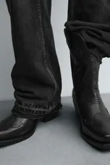 Bota estilo cowboy de caña media, confeccionada en piel negra con costuras decorativas en la caña y el empeine. Presenta una horma en punta, tacón ancho y tiradores laterales.