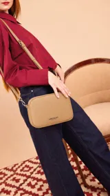 Bandolera pequeña de diseño compacto en cuero sintético color beige, con correa ajustable del mismo material y cierre superior. Presenta el logo de la marca grabado en la parte frontal.