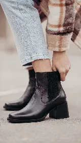 Bota tipo Chelsea de cuero negro, con paneles laterales elásticos y taco cuadrado de 6 cm con acabado de madera. Diseño clásico con puntera redondeada y tirador trasero.