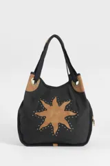 Bolso oversize negro de canvas con diseño de sol en eco-cuero marrón aplicado en el frente, bordeado con tachas metálicas. Cuenta con asas de hombro y detalles de herrajes en tono bronce.