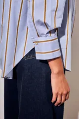 Camisa de corte oversize con estampado de rayas verticales en tonos azul claro y mostaza. Presenta cuello clásico, cierre frontal con botones y mangas largas con puños abotonados.