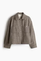 Chaqueta de tejido grueso con estampado pata de gallo en tonos beige, marrón y negro. Presenta cuello camisero, cierre frontal con botones metálicos plateados, y dos bolsillos de parche con solapa y botón en el pecho. El corte es holgado y ligeramente corto, con mangas largas.