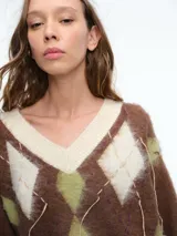 Sweater de tejido de punto con diseño de rombos (argyle) en tonos marrón, verde oliva y crema. Presenta escote en V, mangas largas con puños ajustados y detalles de hilos colgantes decorativos en el frente que le otorgan un aspecto desgastado y moderno.