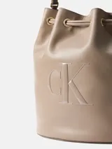 Cartera tipo bucket de Calvin Klein en color beige, confeccionada en poliuretano con asa superior, cierre con cordón ajustable y monograma de la marca grabado en el frente.