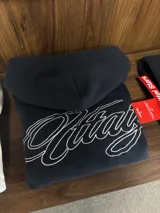 Hoodie negro con capucha y estampa gráfica blanca en la espalda con diseño tipográfico cursivo. Presenta puños y ruedo acanalados.