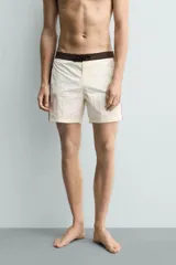 Short de baño blanco marfil con cintura marrón y cordón ajustable del mismo color. Presenta franjas laterales en tono azul marino con finos bordes rojizos y bolsillos laterales.