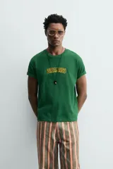 Camiseta de corte relajado, color verde, con cuello redondo y manga corta. Presenta un texto bordado en relieve con letras amarillas y contorno oscuro en el centro del pecho.