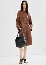 Chaqueta larga de cuero sintético marrón con efecto charol, solapas clásicas, cierre frontal oculto y cinturón ajustable con hebilla del mismo material. Cuenta con bolsillos laterales y mangas largas con trabillas en los puños.