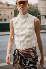 Chaleco de punto en color crudo con cuello subido y manga sisa. Presenta un diseño de tejido con estructura de cables gruesos en el frente y acabados acanalados en el cuello y el bajo.