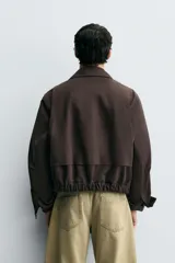 Cazadora cropped fit color marrón oscuro, confeccionada en tejido de algodón. Presenta cuello solapa, manga larga con puño abotonado, bolsillos de vivo en la cadera y un bolsillo interior. El bajo tiene elástico en la espalda y el cierre es frontal con cremallera.