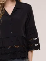 Camisa negra de poliéster con cuello camisero, escote en V y cierre frontal de botones. Cuenta con mangas anchas de corte acampanado y detalles de bordado suizo calado en las mangas y en el ruedo inferior.