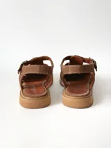 Sandalias estilo cangrejera color marrón, confeccionadas en cuero con diseño trenzado en la parte delantera, tira ajustable con hebilla metálica en el tobillo y suela plana.