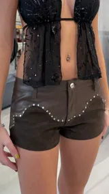Short marrón de tiro bajo confeccionado en cuero sintético y gamuza, con detalles de tachas metálicas en las costuras y diseño de bolsillos traseros con cortes curvos.
