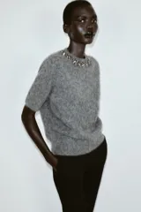 Jersey de punto de manga corta en color gris jaspeado, con cuello redondo decorado con abalorios joya y cierre de botón en la espalda.