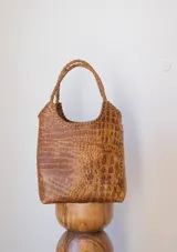 Bolso tipo matera de cuero con textura de reptil, color marrón. Presenta asas trenzadas y costuras artesanales en los bordes.
